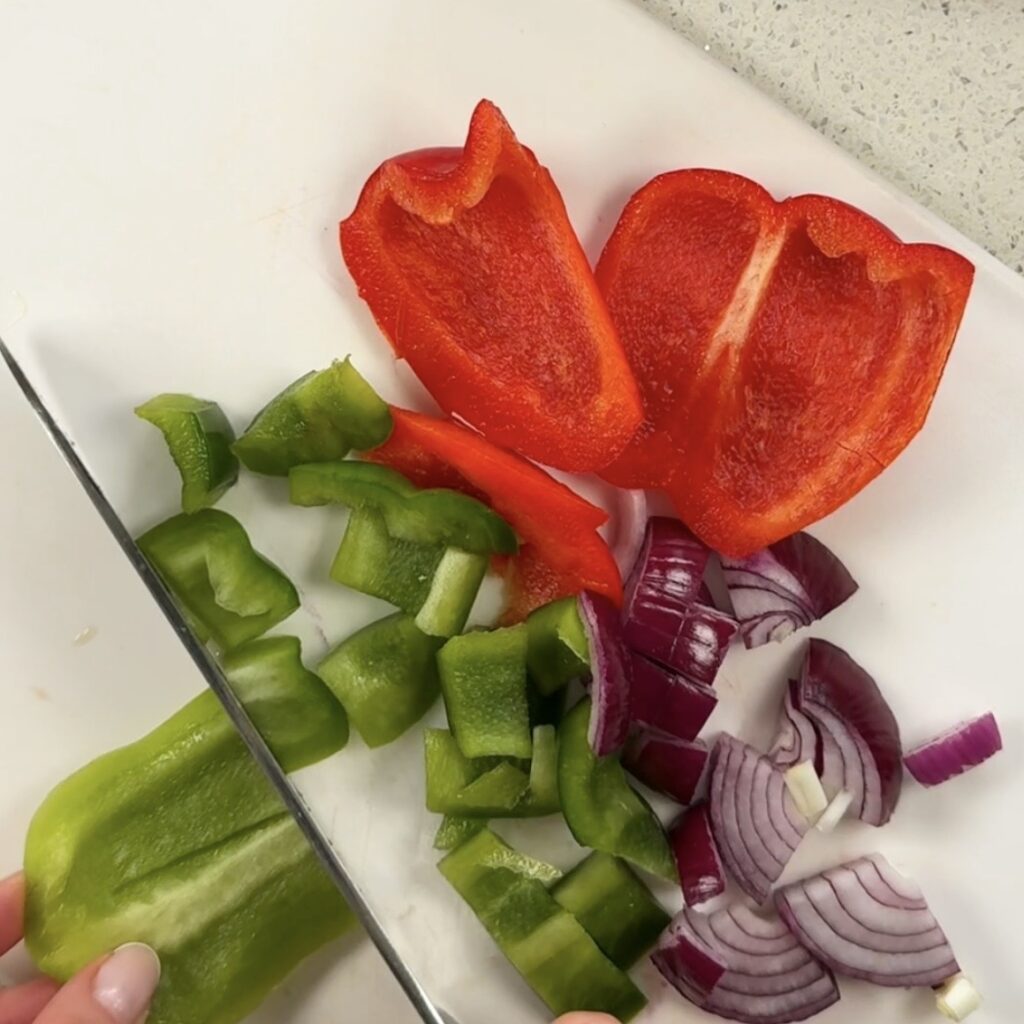 Red pepper, green pepper and red onion being chopped on a white chopping board