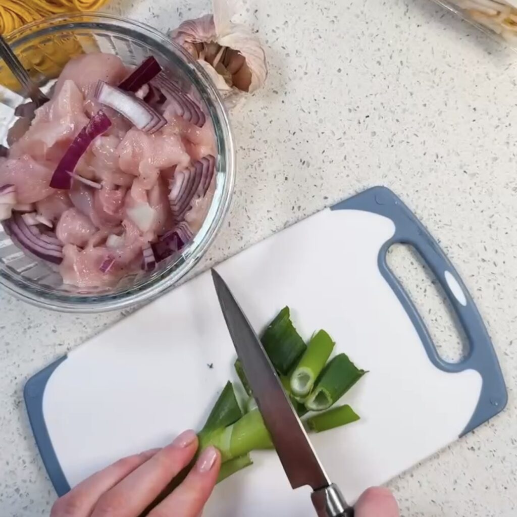 Sliced chicken breast and red onion in a glass bowl with chopped spring onions on a chopping board