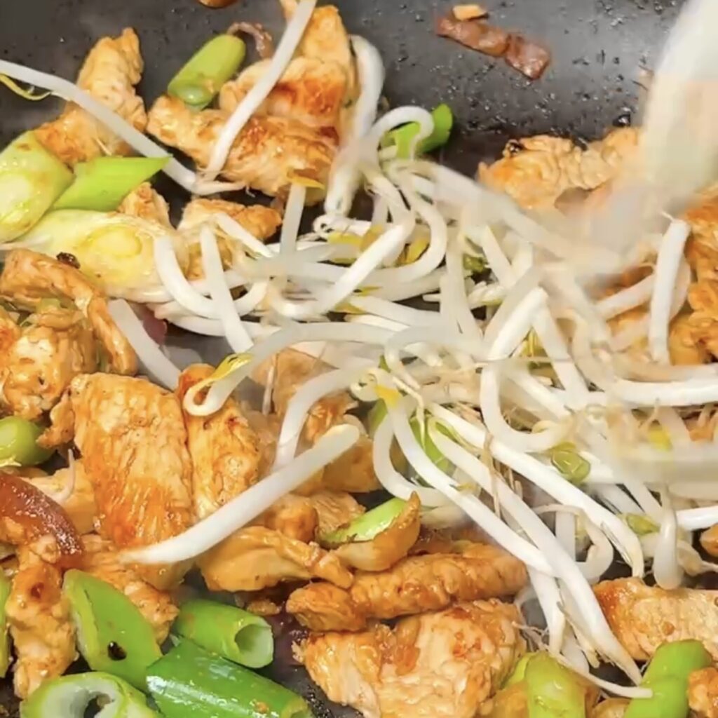 Bean sprouts being added to chicken and spring onions in a wok