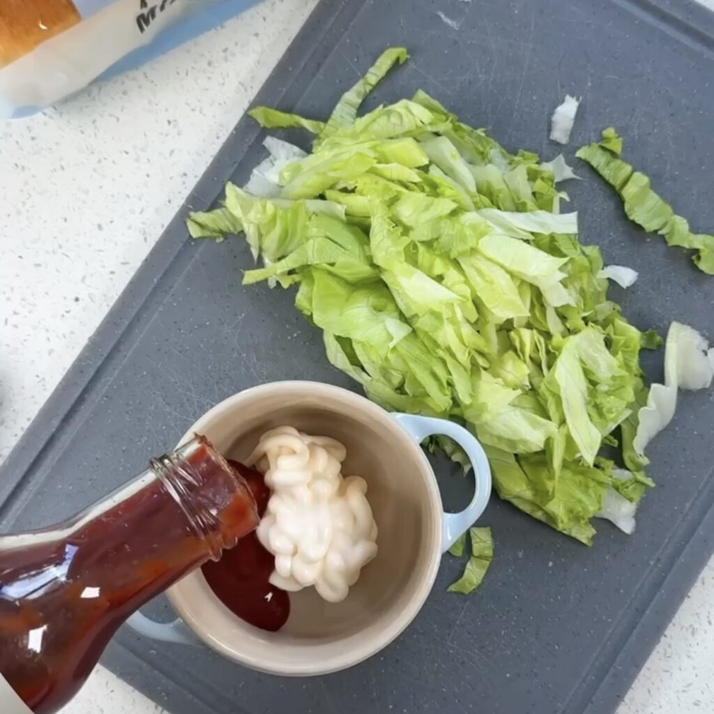 Spicy ketchup being poured into a bowl of mayonnaise next to shredded iceberg lettuce on a chopping board
