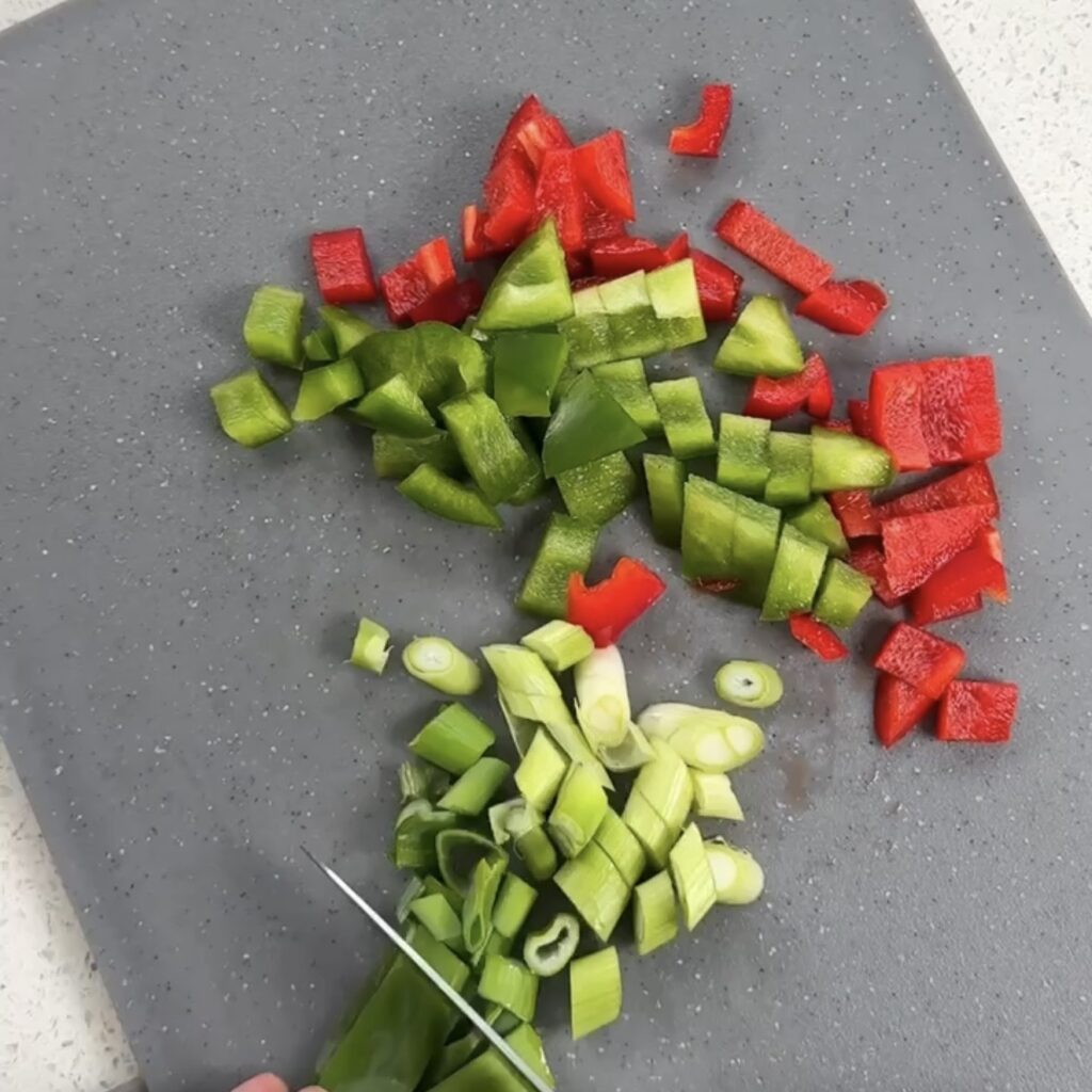 Red pepper, green pepper and spring onion being chopped on a grey chopping board
