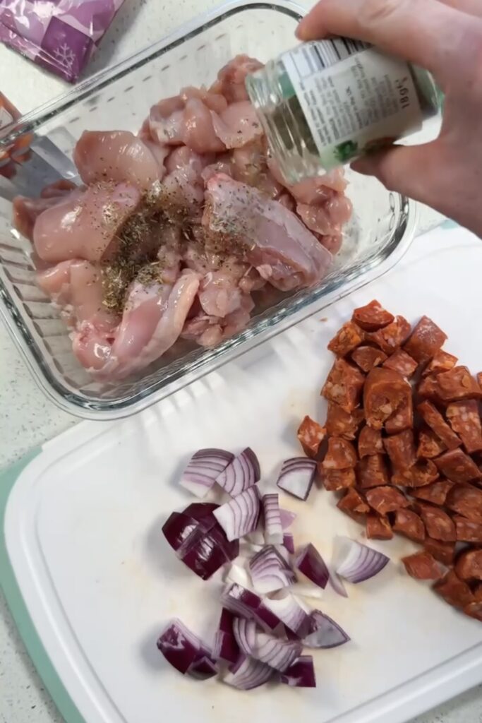 Diced chicken thighs being seasoned with Italian herbs in a glass dish, with chopped red onion and chorizo on a chopping board