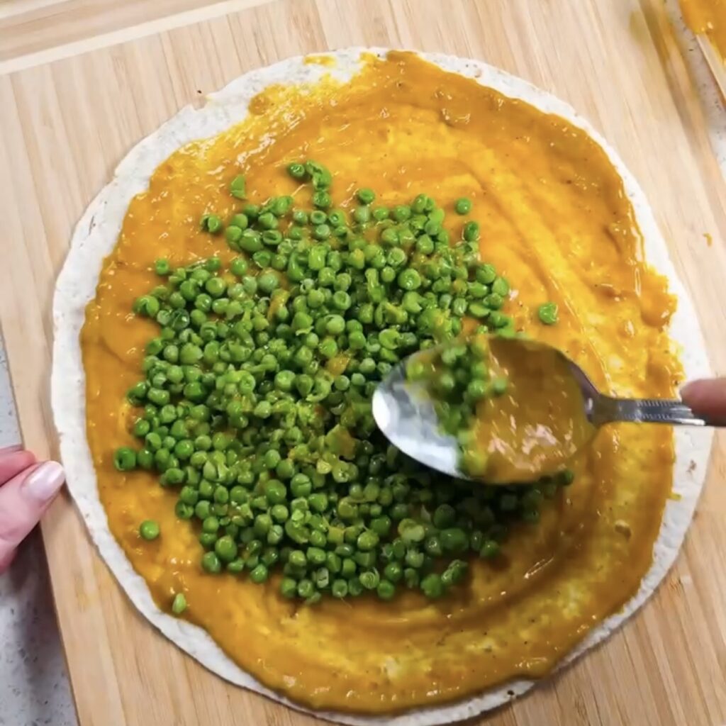 Crushed peas being spooned onto a wrap spread with curry sauce