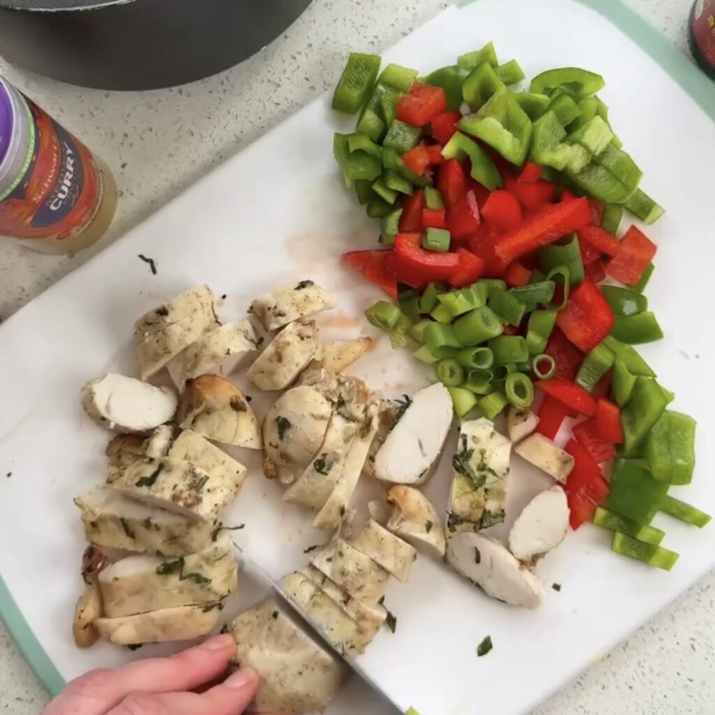 Sliced cooked chicken breast and chopped red and green pepper and spring onion on a chopping board