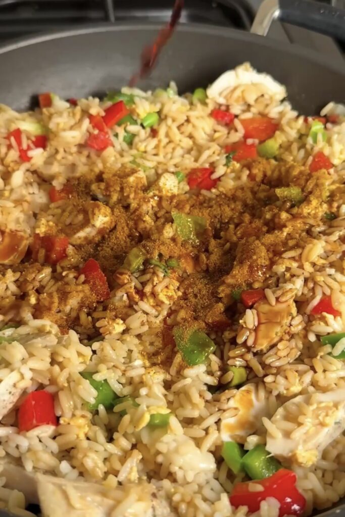Curry powder being sprinkled over chicken fried rice with peppers in a pan