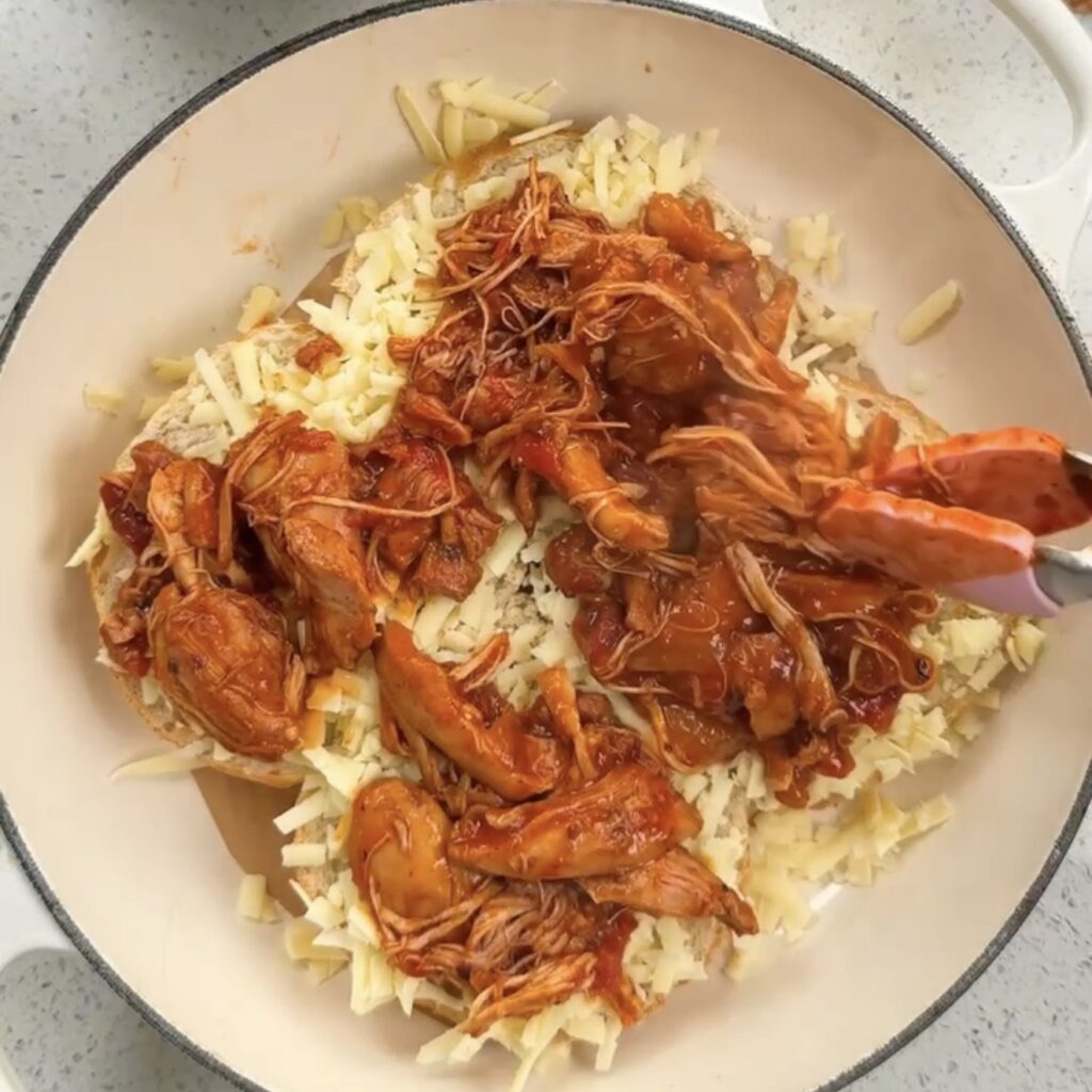 Shredded BBQ hunter's chicken being added to crusty rolls lined with grated cheese using tongs